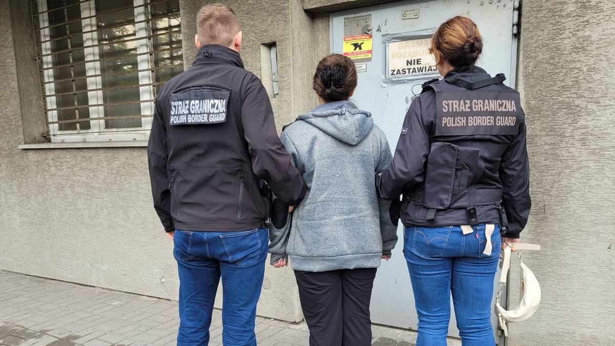 Dwóch funkcjonariuszy Straży Granicznej w kurtkach z napisem "STRAŻ GRANICZNA" odprowadza zatrzymaną osobę w szarej bluzie.