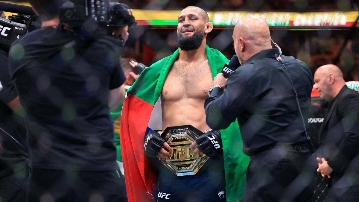 Zwycięzca walki UFC, Khamzat Chimaev, trzyma pas mistrzowski i jest owijany flagą narodową.