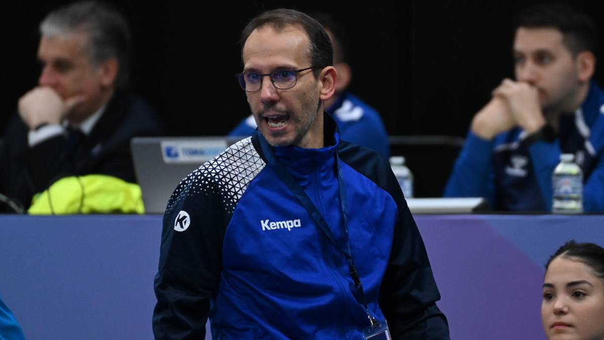 Mężczyzna w niebiesko-białej kurtce sportowej i okularach, mówiący do przodu.