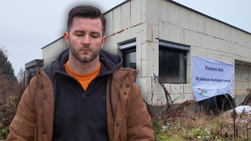 Mężczyzna w brązowej kurtce stoi przed niedokończonym domem. Na budynku wisi baner z napisem "Kupujesz dom. W pakiecie dostajesz dom".