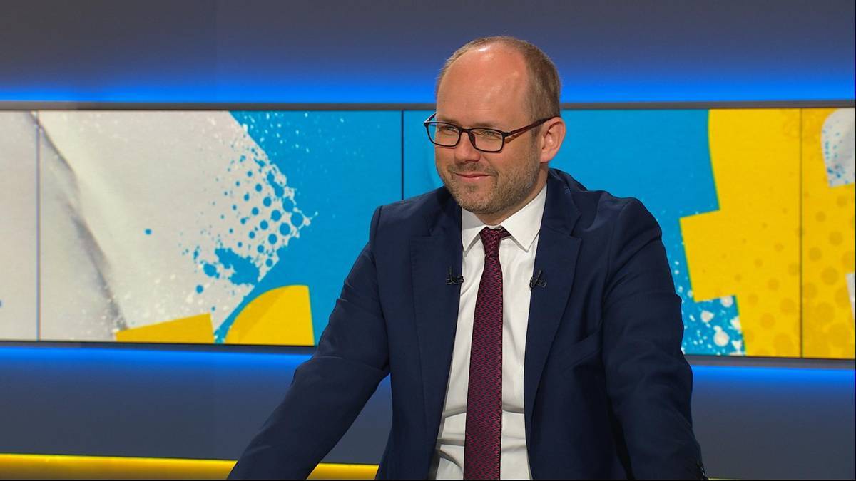 Mężczyzna w okularach i garniturze z krawatem uśmiecha się przed niebiesko-żółtym tłem.