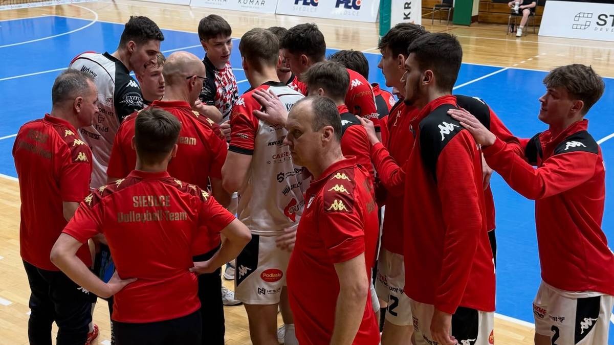 Drużyna siatkarzy w czerwonych strojach zgromadzona w kręgu w hali sportowej.