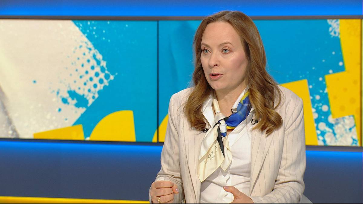 Kobieta w jasnej marynarce, z apaszką na szyi, siedzi i m&oacute;wi, na tle jasnoniebieskich ekran&oacute;w z abstrakcyjnymi wzorami w kolorach białym, ż&oacute;łtym i niebieskim.