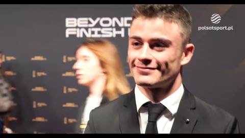 Mężczyzna w garniturze i krawacie z lekkim uśmiechem na twarzy, stoi na tle napisu "BEYOND FINISH" i logo Polsat Sport.