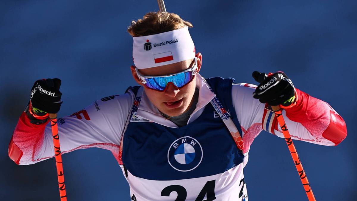Biathlonista w stroju reprezentacji Polski z numerem startowym 24 na piersi i czerwonych spodniach narciarskich, biegnący na nartach po ośnieżonym stoku z kijami narciarskimi w dłoniach, z widoczną latarnią w tle.