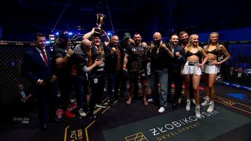 Grupa zawodników MMA pozuje do zdjęcia z trofeum, świętując zwycięstwo.