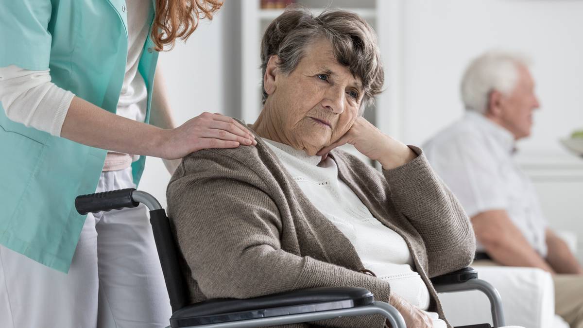 Starsza kobieta siedzi na w&oacute;zku inwalidzkim, z ręką na biodrze i zamyślonym wyrazem twarzy. Obok niej stoi inna osoba, kt&oacute;ra kładzie jej dłoń na ramieniu.