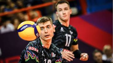 Dw&oacute;ch siatkarzy w czarnych strojach sportowych na boisku. Jeden z nich, z numerem 16 na koszulce, jest na pierwszym planie i skupiony na piłce do siatk&oacute;wki, kt&oacute;ra znajduje się w powietrzu obok jego głowy. Drugi siatkarz, z numerem 17, stoi za nim.