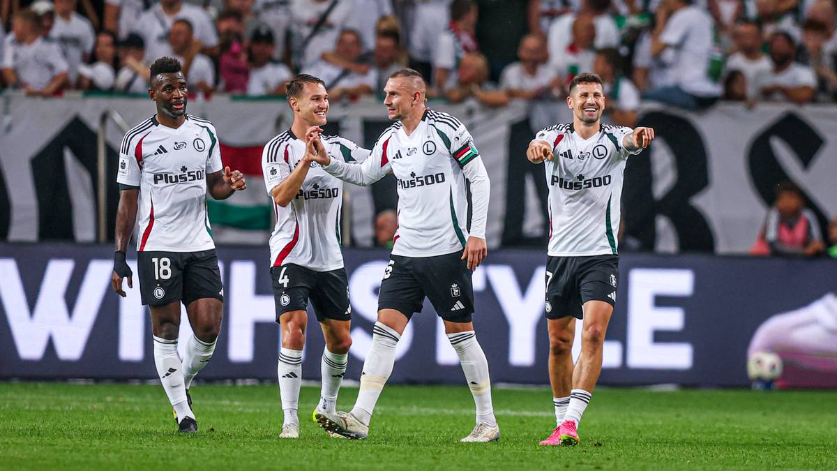 Czterech piłkarzy Legii Warszawa świętuje zwycięstwo na murawie stadionu. Zawodnicy ubrani są w białe koszulki z czarnymi elementami i numerami na plecach. Tworzą radosny zbi&oacute;r, wykonując gesty radości.