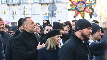 Tłum ludzi w zimowych ubraniach, część os&oacute;b w czapkach, w tle budynek i ozdoba przypominająca gwiazdę.