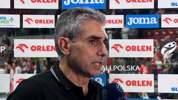 Portret mężczyzny z siwiejącymi włosami, m&oacute;wiącego do mikrofonu, na tle baner&oacute;w sponsor&oacute;w.