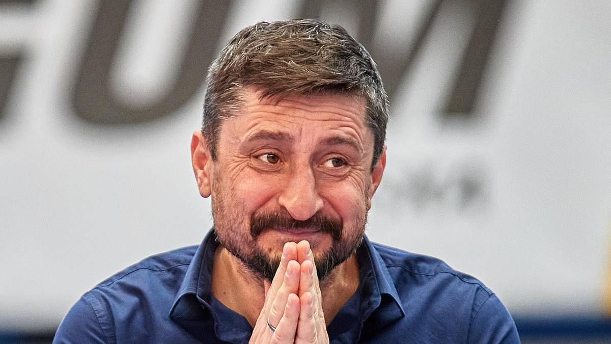 Trener siatkówki w ciemnej koszuli, z zarostem, z dłońmi złożonymi w geście prośby, uśmiecha się.