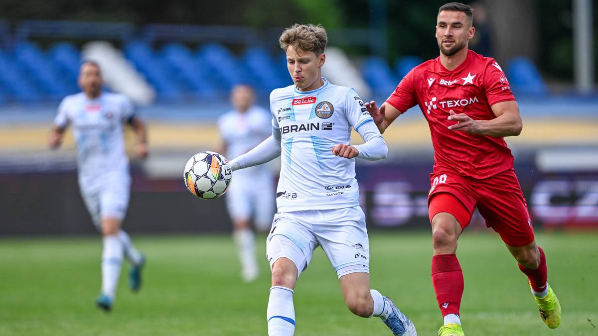 Piłkarz w białej koszulce prowadzi piłkę, a za nim biegnie zawodnik w czerwonej koszulce.