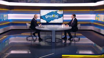 Dw&oacute;ch mężczyzn siedzi naprzeciwko siebie przy stole w studiu telewizyjnym, na kt&oacute;rym wyświetlane jest logo "graffiti".