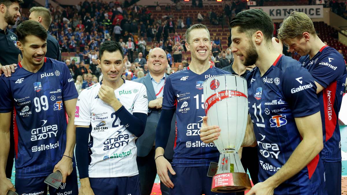 Pięciu siatkarzy w ciemnych strojach sportowych, jeden z nich trzyma trofeum.