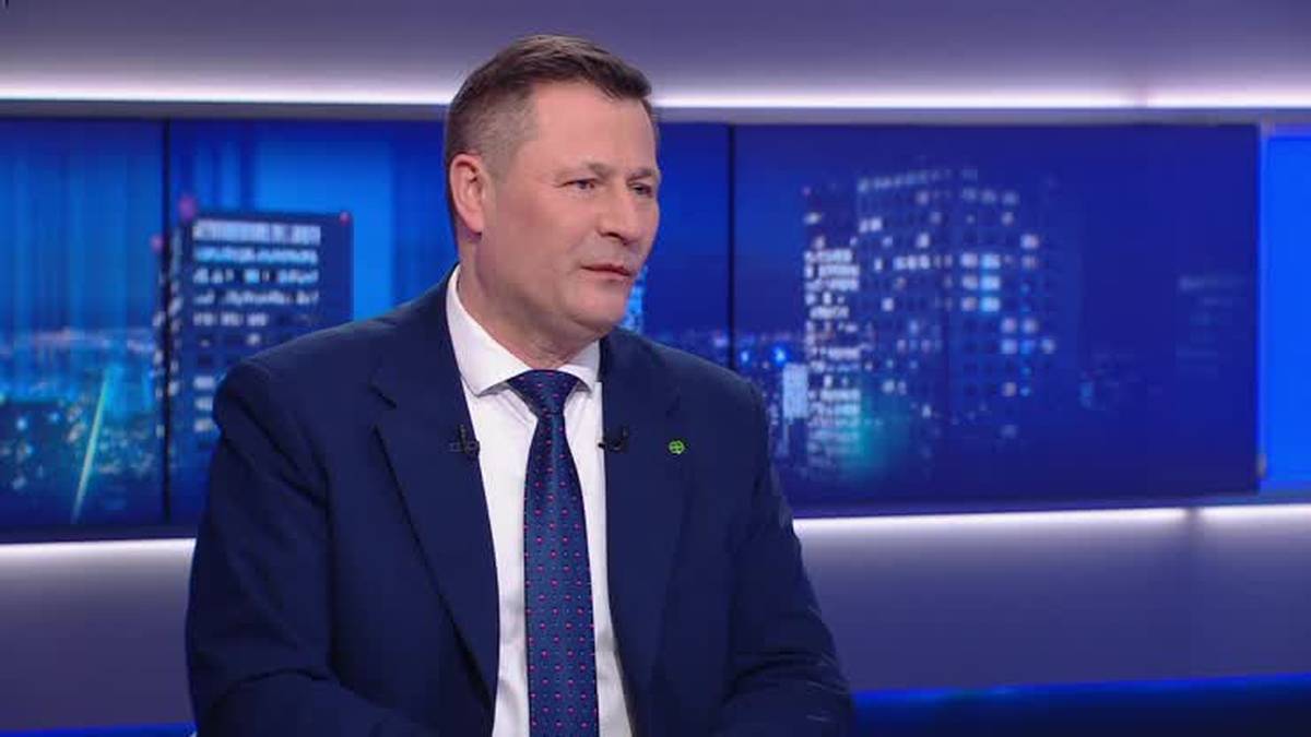 Krzysztof Paszyk w garniturze i krawacie siedzi i m&oacute;wi.