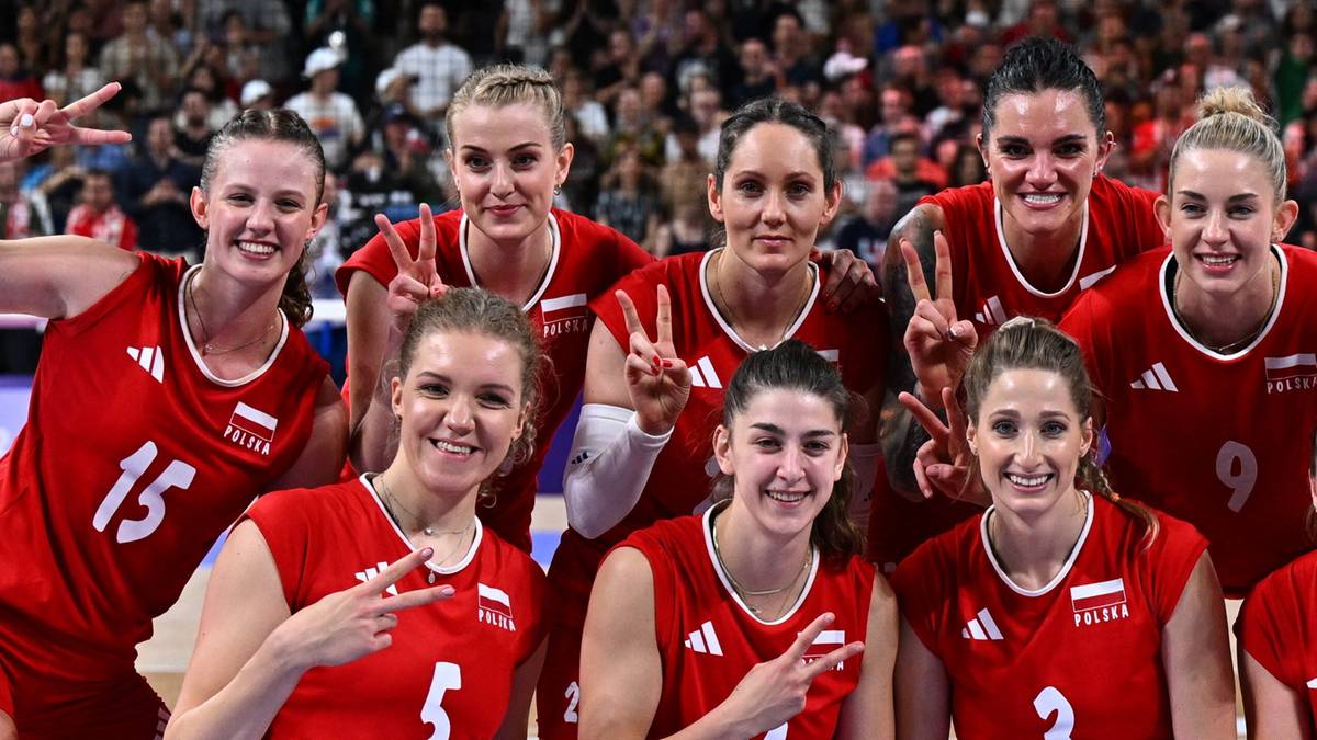 Grupa polskich siatkarek w czerwonych strojach sportowych, z numerami na koszulkach, pozuje do zdjęcia z uśmiechami na twarzach.