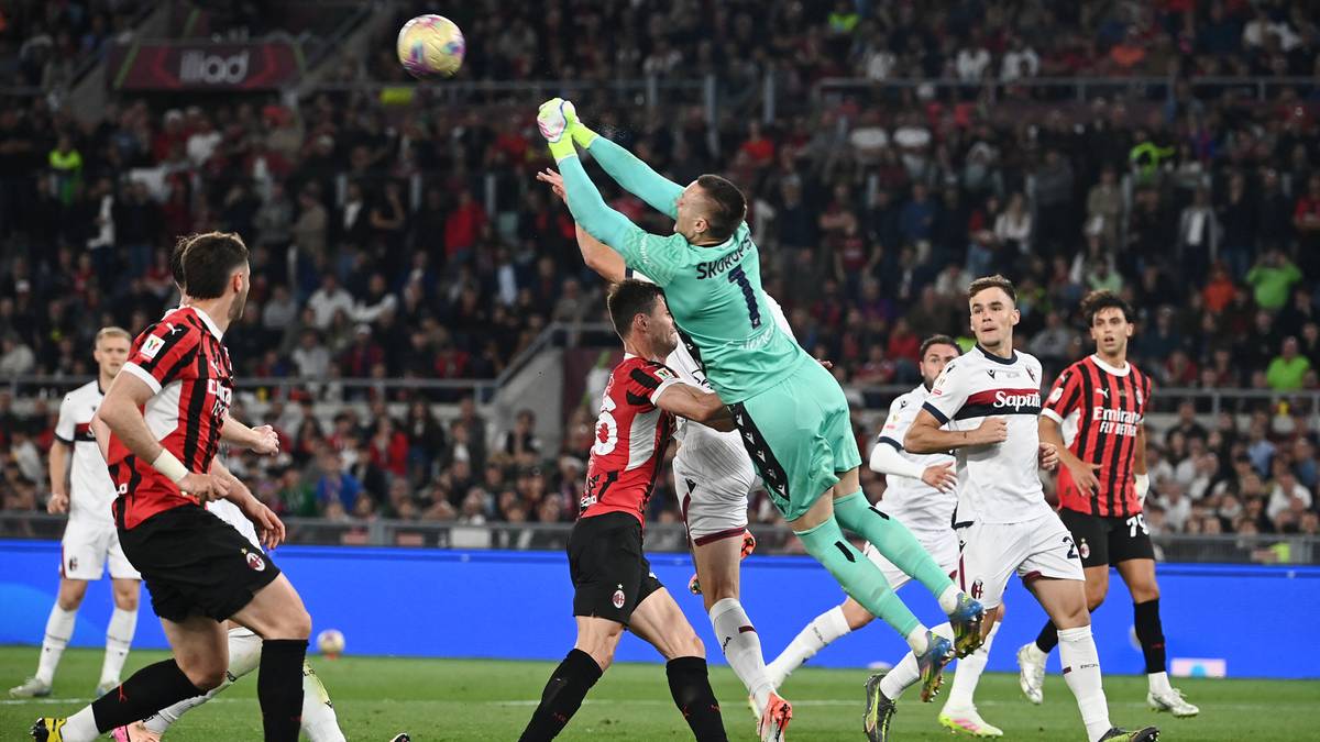 Bramkarz w niebieskim stroju wyskakuje, aby obronić piłkę lecącą w powietrzu podczas meczu piłki nożnej.