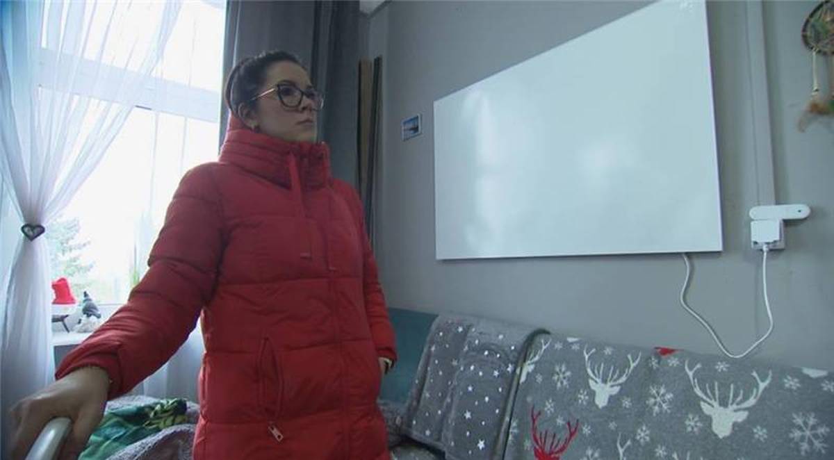 Kobieta w czerwonej kurtce stoi w pomieszczeniu z zamontowanym na ścianie białym panelem grzewczym.