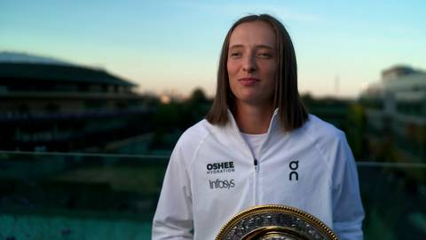 Iga Świątek, polska tenisistka, w białej bluzie sportowej z logo OSHЕE i Infosys, trzyma trofeum na tle budynków i nieba o zachodzie słońca.