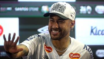 Uśmiechnięty mężczyzna, piłkarz Neymar, w białej koszulce sportowej i czapce, z uniesioną dłonią.