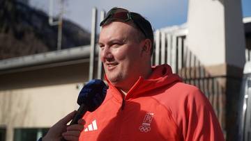 Mariusz Hluchnik w pomarańczowej bluzie z kapturem, udziela wywiadu z mikrofonem przed budynkiem.