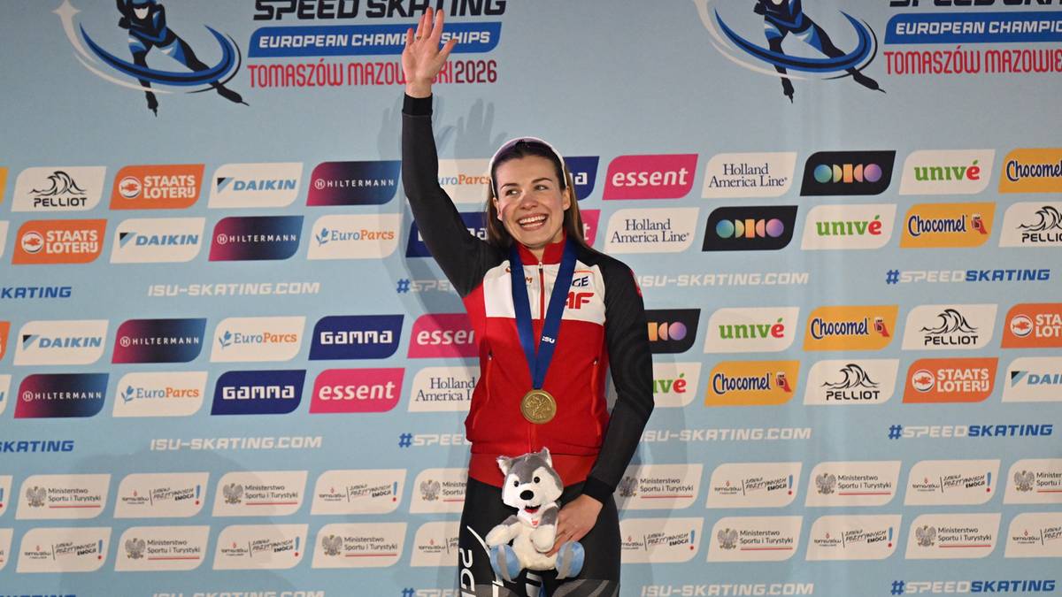 Uśmiechnięta sportsmenka z medalem na szyi i pluszową maskotką pozuje na tle ścianki sponsorskiej.