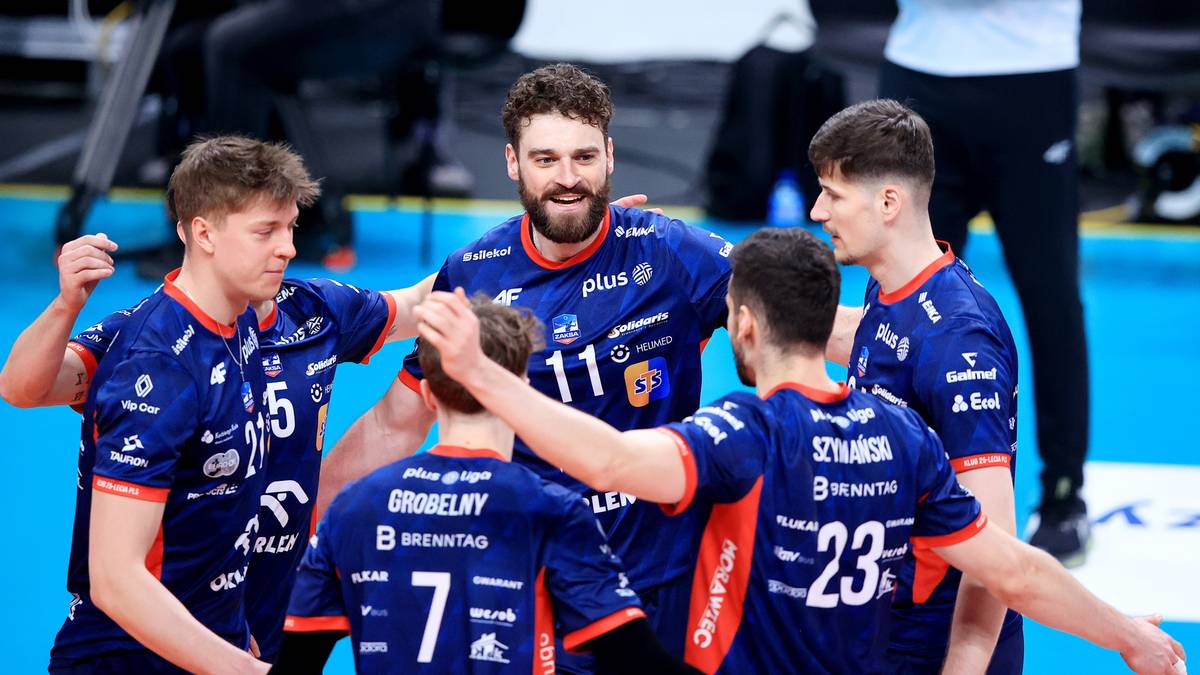 Sześciu siatkarzy w granatowych strojach z numerami na plecach, stojących blisko siebie i rozmawiających podczas meczu siatk&oacute;wki.