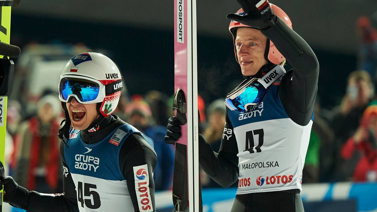 Dw&oacute;ch skoczk&oacute;w narciarskich w kaskach i goglach, patrzących w g&oacute;rę z radosnymi wyrazami twarzy.