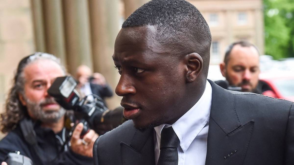 Mężczyzna o ciemnej sk&oacute;rze w eleganckim garniturze, z uwagą wpatrzony w bok. Za nim widać zamazane postacie i aparat fotograficzny.