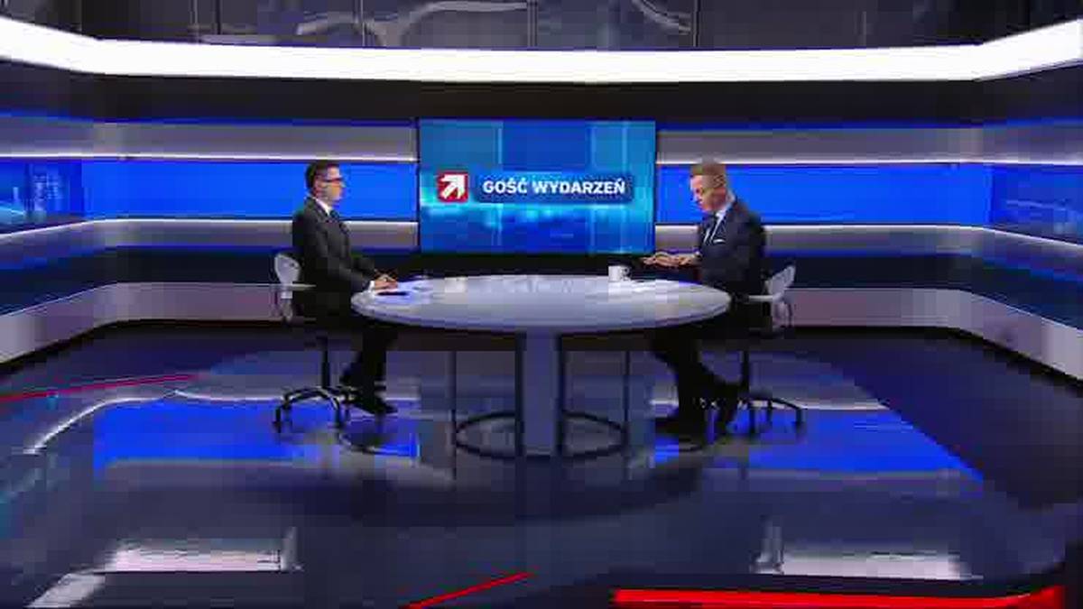 Dw&oacute;ch mężczyzn w garniturach siedzi naprzeciwko siebie przy owalnym stole w studiu telewizyjnym. Na ekranie za nimi wyświetla się logo programu "Gość Wydarzeń".