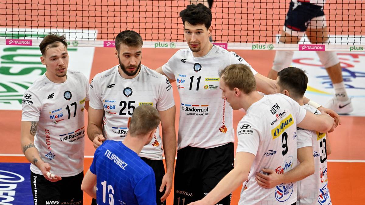 Grupa mężczyzn w strojach siatkarskich stoi na boisku, rozmawiając. Dwóch zawodników obejmuje się nawzajem.