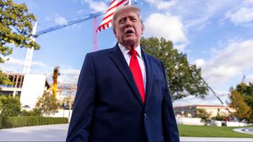 Donald Trump w granatowym garniturze i czerwonym krawacie na tle błękitnego nieba i amerykańskiej flagi.