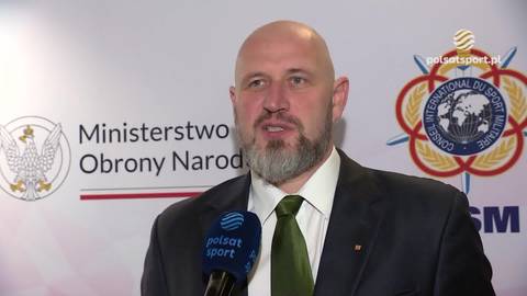 Tomasz Majewski w garniturze z krawatem, udziela wywiadu przed ścianką z logotypami Ministerstwa Obrony Narodowej i Międzynarodowej Rady Sportu Wojskowego. Przed nim stoi mikrofon Polsat Sport.