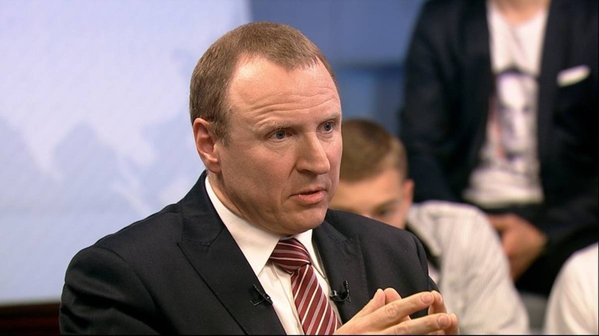 Mężczyzna w garniturze i krawacie mówi, jego ręce są splecione przed nim.