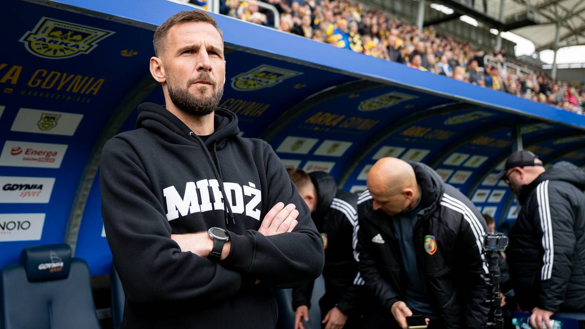 Mężczyzna w czarnej bluzie z kapturem stoi z założonymi rękami, patrząc przed siebie z poważną miną. Za nim widać ławkę rezerwowych i trybuny stadionu z kibicami.