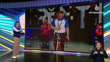 Kobieta po prawej stronie z szeroko otwartymi ustami i oczami, ubrana w ciemną bluzę z kapturem, wydaje się być zaskoczona lub podekscytowana. Na dużym ekranie w tle widać mężczyznę w niebiesko-białej koszulce sportowej, siedzącego na specjalistycznym rowerze. Obok niego stoi mężczyzna w kamizelce i okularach, trzymający niebieską barierkę. W tle ekranu znajdują się inne osoby w mundurach lub strojach sportowych.