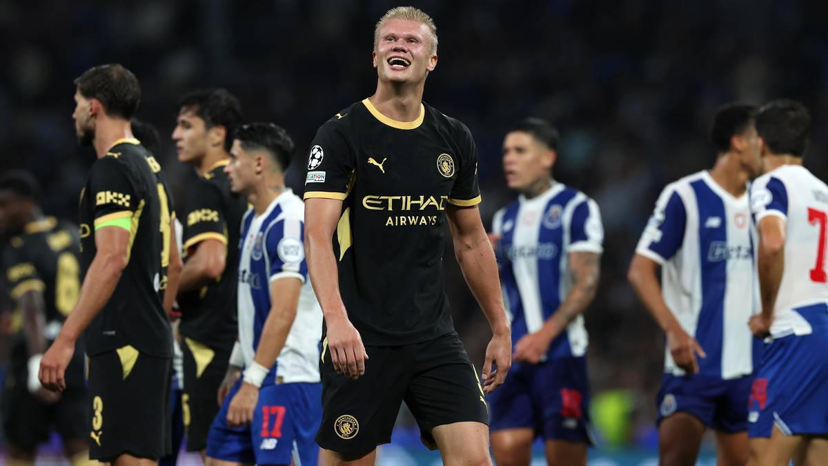 Erling Haaland w czarnej koszulce Manchesteru City z ż&oacute;łtymi detalami patrzy w g&oacute;rę z uśmiechem. Wok&oacute;ł niego stoją inni piłkarze w niebiesko-białych strojach.