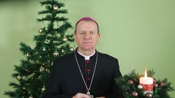 Abp Tadeusz Wojda w stroju duchownym stoi przed choinką i zapaloną świecą.