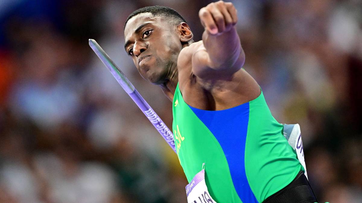 Atletyczny mężczyzna w zielonej koszulce z niebieskimi wstawkami wykonuje rzut oszczepem. Jego twarz wyraża determinację i skupienie.