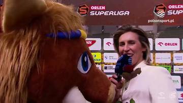Maskotka w stroju konia zbliża się do mikrofonu, przerywając wywiad dziennikarki. W tle widoczne są logotypy sportowe.