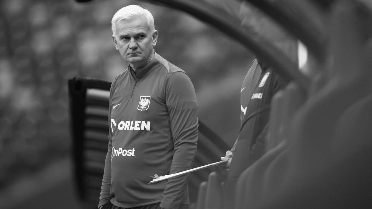 Starszy mężczyzna o siwych włosach, ubrany w sportową bluzę z logo ORLEN, stoi na tle ławki rezerwowych podczas meczu.