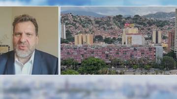 Fragment z wywiadu z mężczyzną w garniturze i widok na zabudowane wzg&oacute;rza Caracas w Wenezueli.