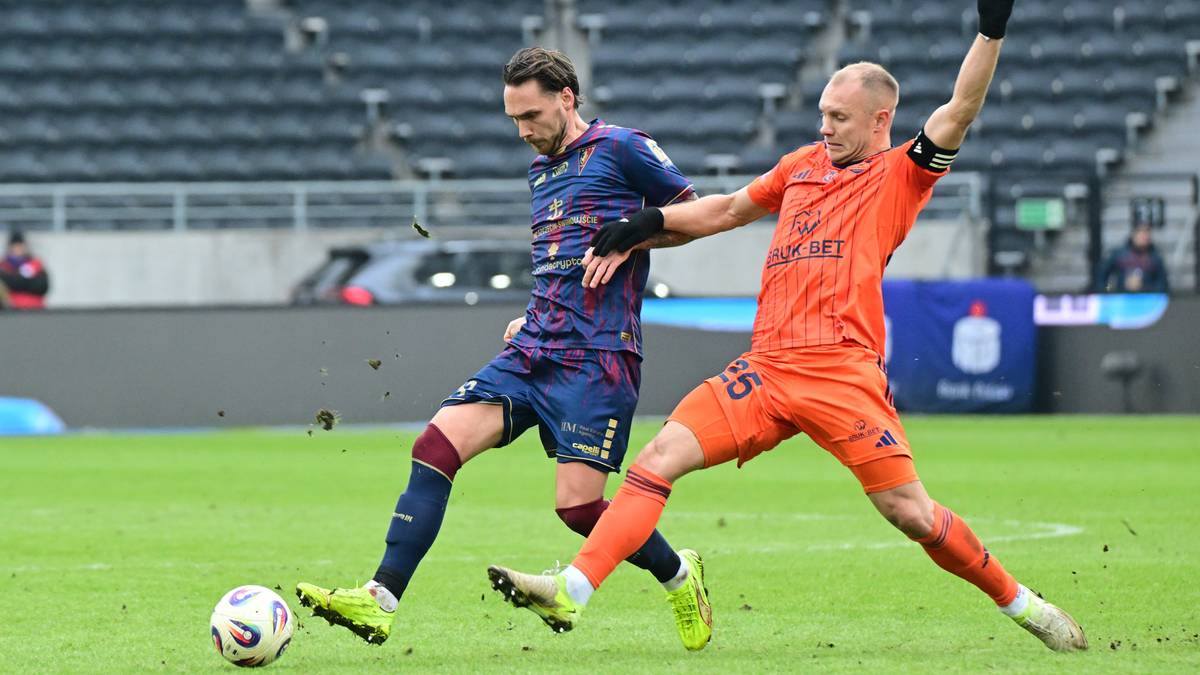 Dw&oacute;ch piłkarzy w akcji na boisku piłkarskim, jeden w niebiesko-czerwonych barwach, drugi w pomarańczowym stroju, pr&oacute;bują przejąć piłkę.