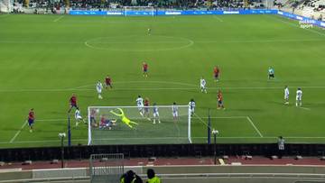 Piłkarz w zielonej bramkarskiej koszulce leży na ziemi, a piłka wpada do bramki po strzale zawodnika w białej koszulce.