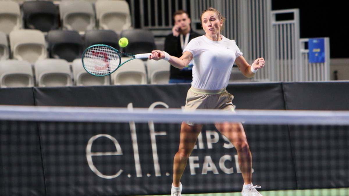 Kobieta w białej koszulce i beżowej spódniczce gra w tenisa, odbija piłkę rakietą.