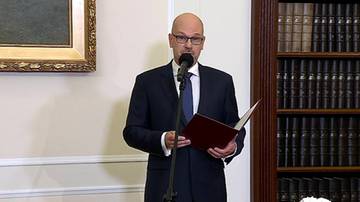 Maciej Berek w garniturze przemawia do mikrofonu, trzymając w rękach dokument na tle regału z książkami.