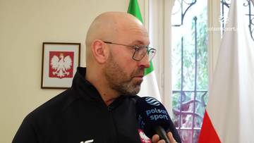 Łysy mężczyzna z brodą i okularami udziela wywiadu. Na jego ramieniu znajduje się mikrofon z logo "Polsat Sport". W tle wiszą dwie flagi Polski i obraz z godłem Polski.