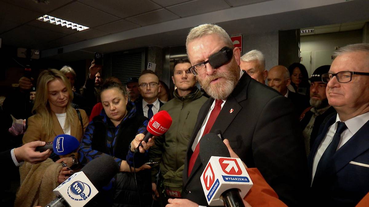 Grzegorz Braun przemawia do dziennikarzy w otoczeniu innych os&oacute;b.