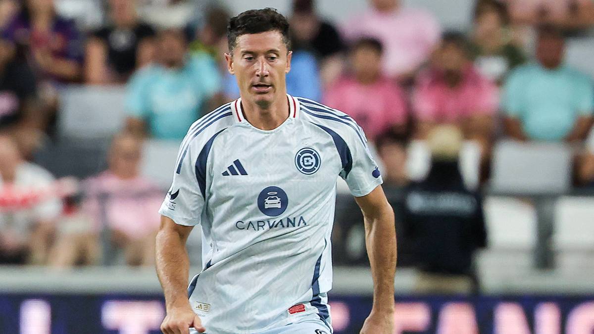 Robert Lewandowski w białej koszulce piłkarskiej z niebieskimi paskami i logo sponsora, na boisku podczas meczu, z niewyraźnymi widzami na trybunach w tle.